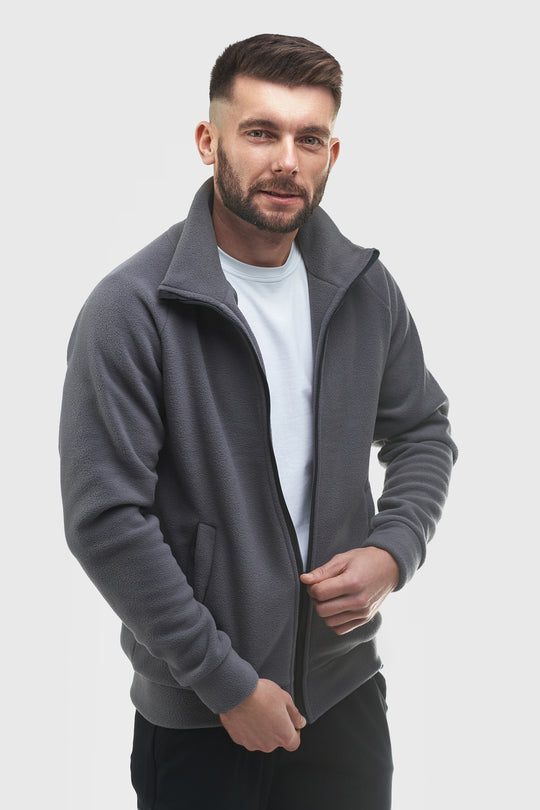 A man wearing a dark gray zip-up fleece jacket over a white t-shirt, standing against a light gray studio background.