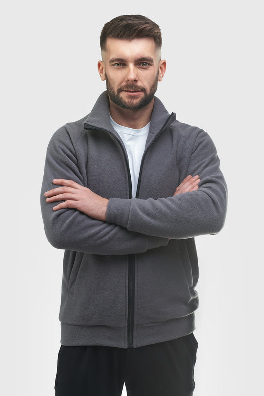 A man with a beard and short hair stands with arms crossed, wearing a dark gray zip-up fleece jacket over a white t-shirt and black pants, against a light gray studio background.