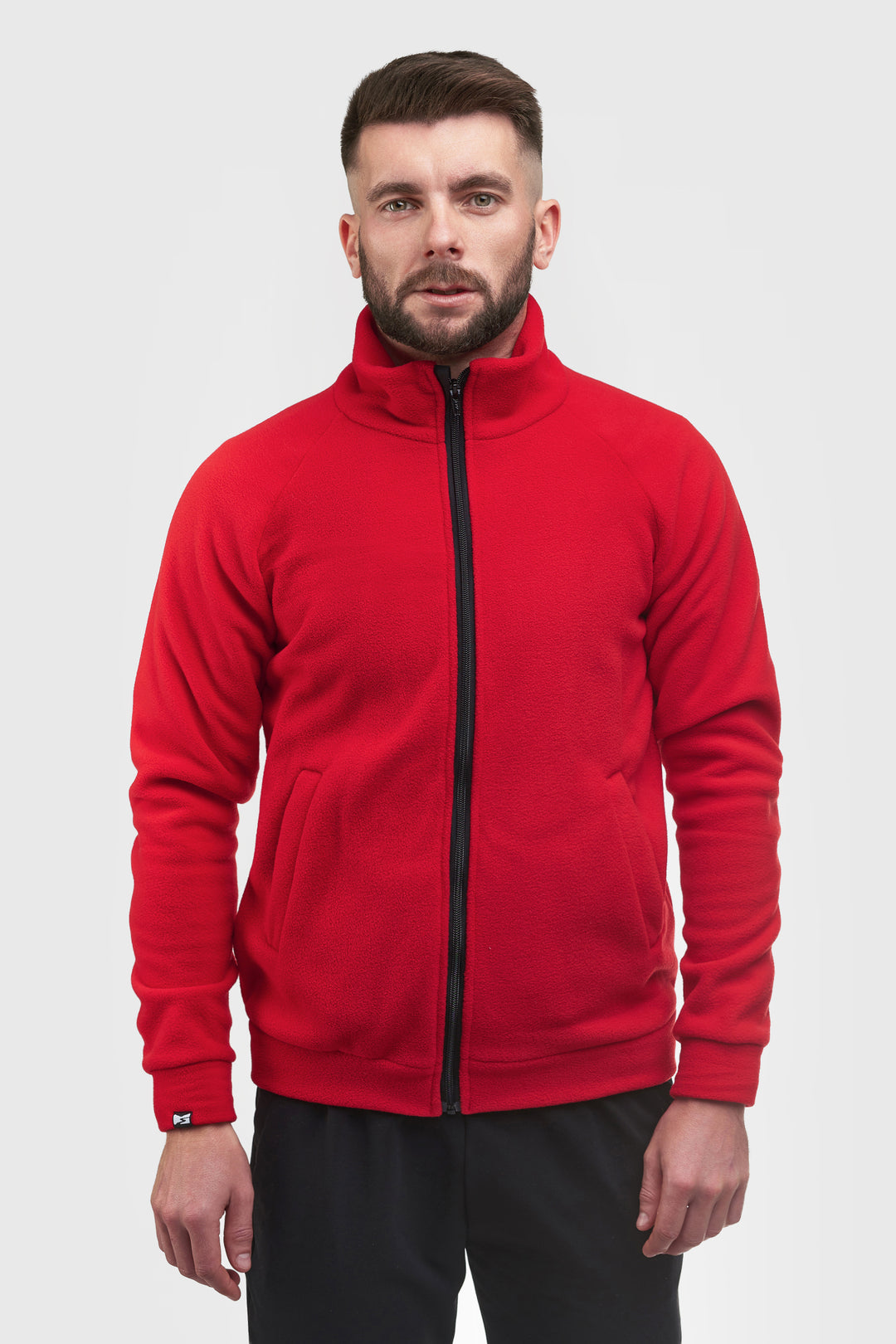 A man wearing a bright red zip-up fleece jacket with a black zipper and black pants, standing against a light gray background.
