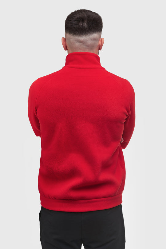 A man viewed from behind wearing a bright red mock-neck or high-collar long-sleeve top with black pants, set against a plain light gray studio background.
