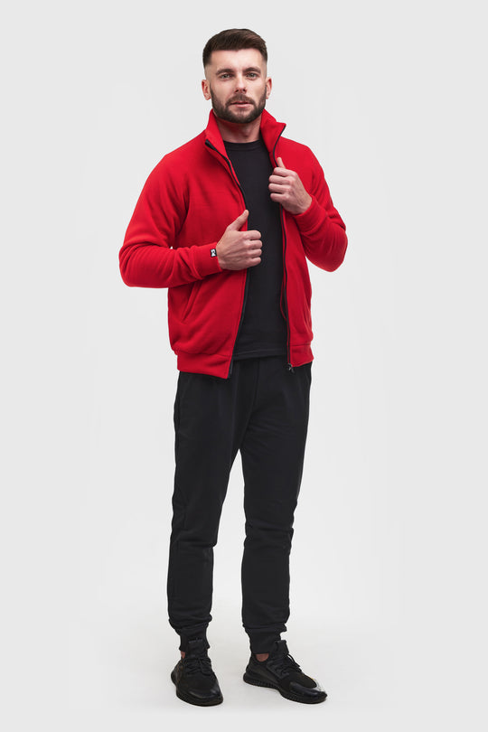 A man modeling a bright red zip-up fleece jacket over a black t-shirt, black joggers, and black sneakers, standing against a light gray studio background.