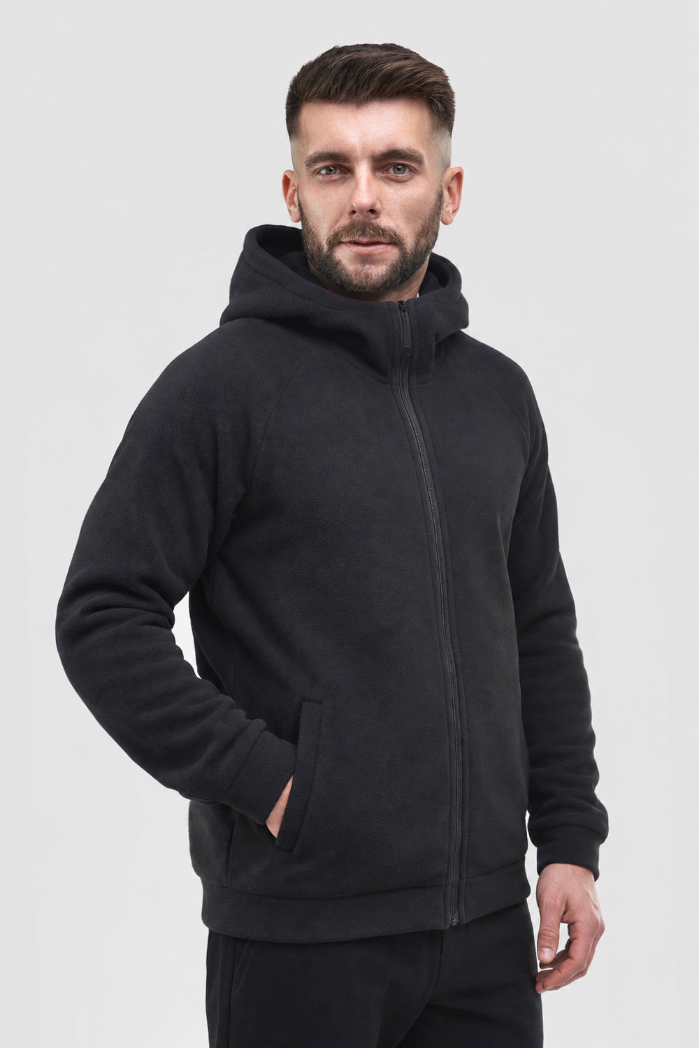 A man wearing a black zip-up fleece hoodie with a high collar and front pockets, standing against a light gray studio background.