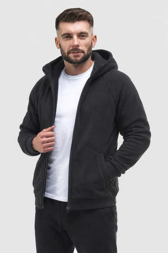 A man wearing a black zip-up hoodie with a kangaroo pocket over a white t-shirt and black pants, standing against a light gray studio background.