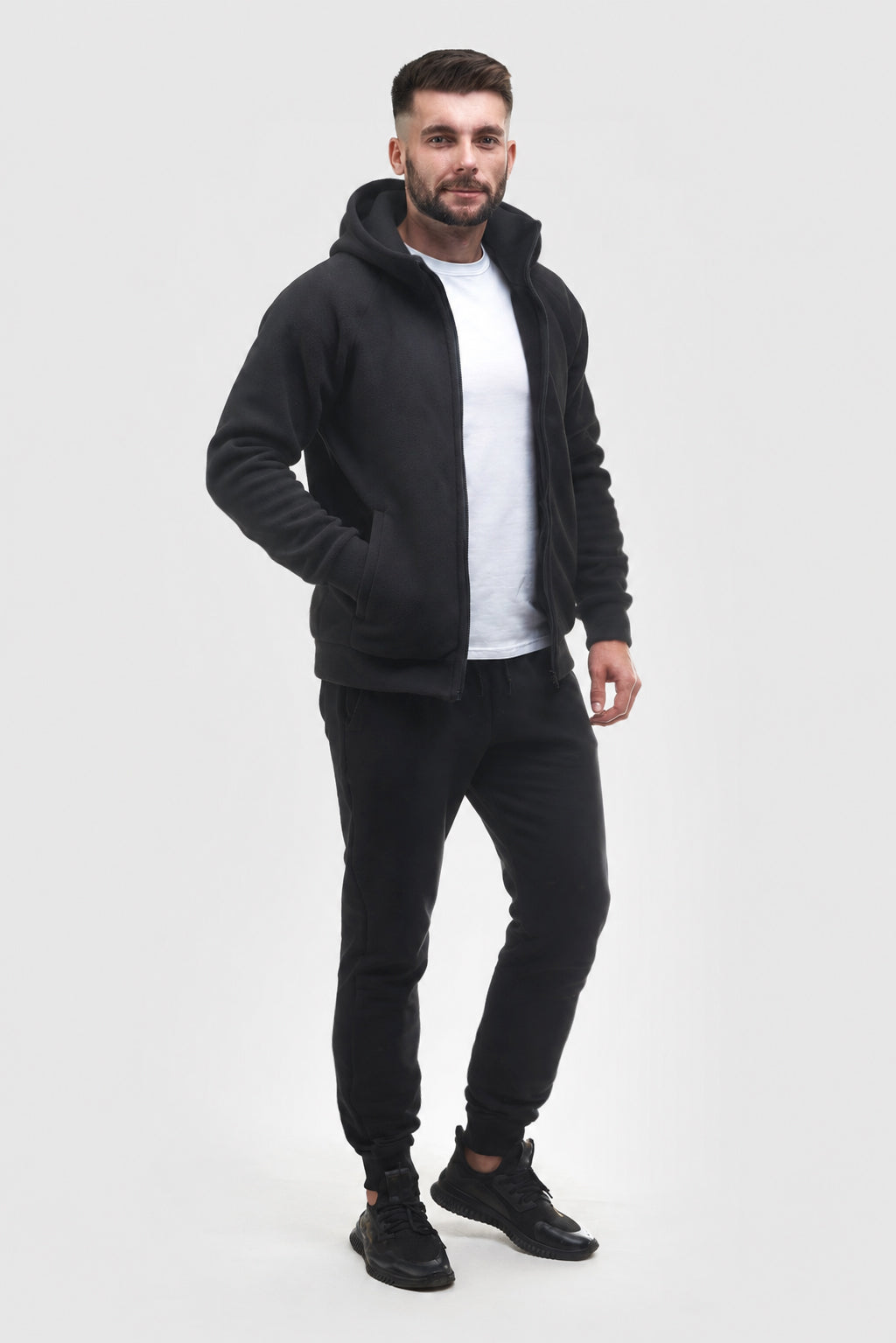 A man modeling a black zip-up hoodie with a hood, worn over a white t-shirt and black joggers, standing against a light gray studio background.