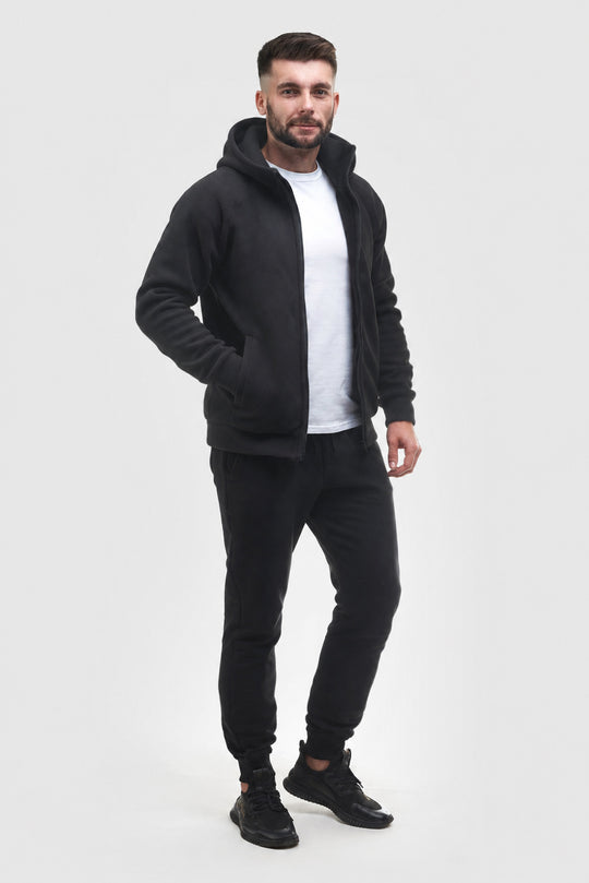 A man modeling a black zip-up hoodie with a hood, worn over a white t-shirt and black joggers, standing against a light gray studio background.