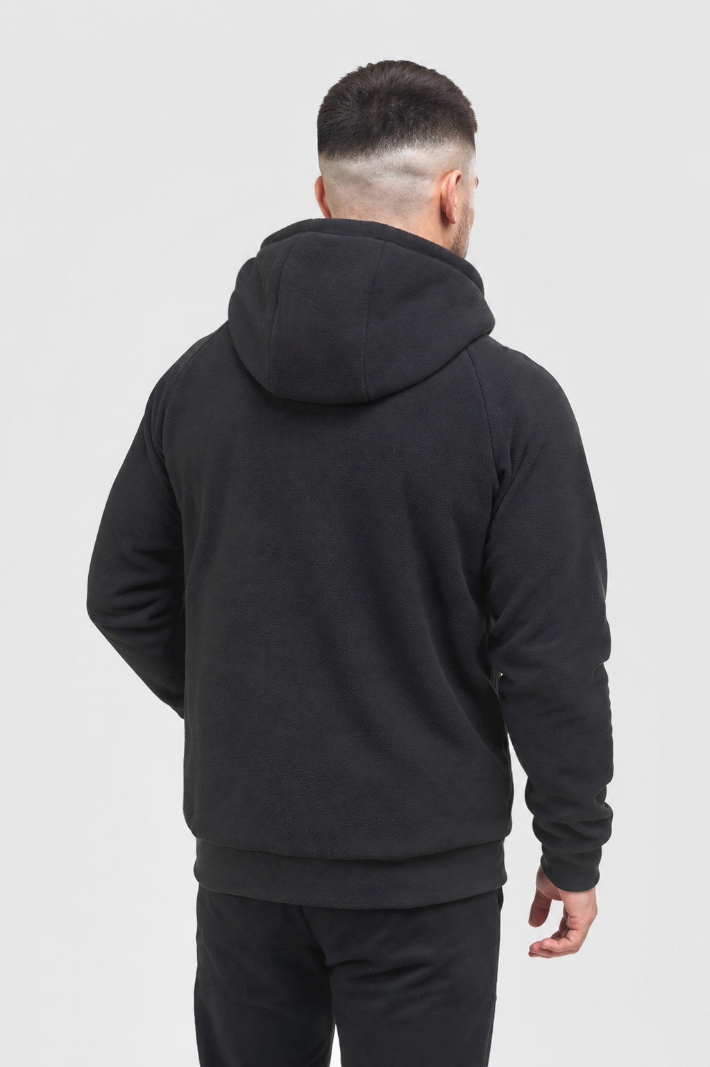 A man viewed from behind wearing a black pullover hoodie and black pants against a light gray studio background.