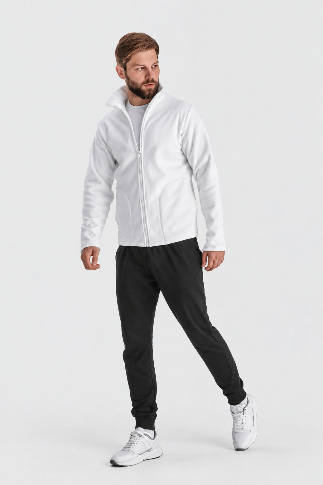 A man modeling a white zip-up fleece jacket, black sweatpants, and white sneakers against a light gray studio background.