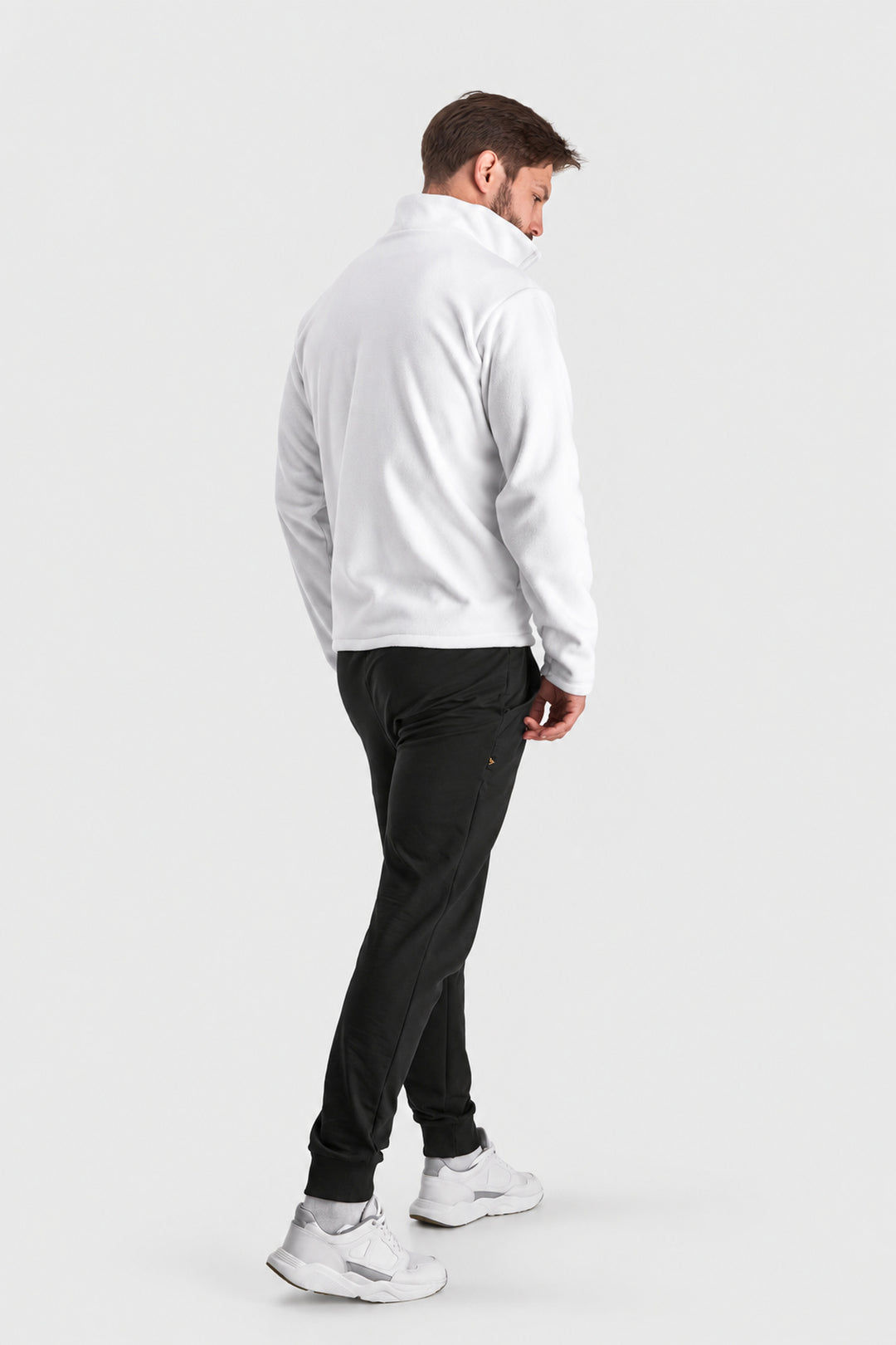 A man shown from the back wearing a white quarter-zip pullover, black jogger pants, and white sneakers, standing against a light gray studio background.