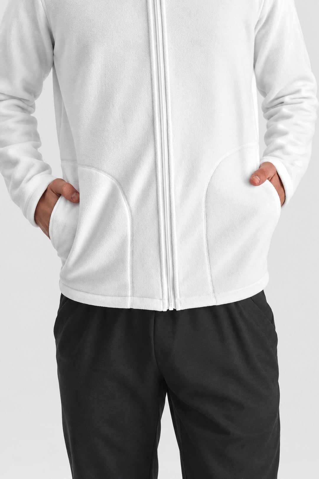 A close-up of a person wearing a white zip-up fleece jacket with hand pockets, paired with black pants, against a light gray studio background.