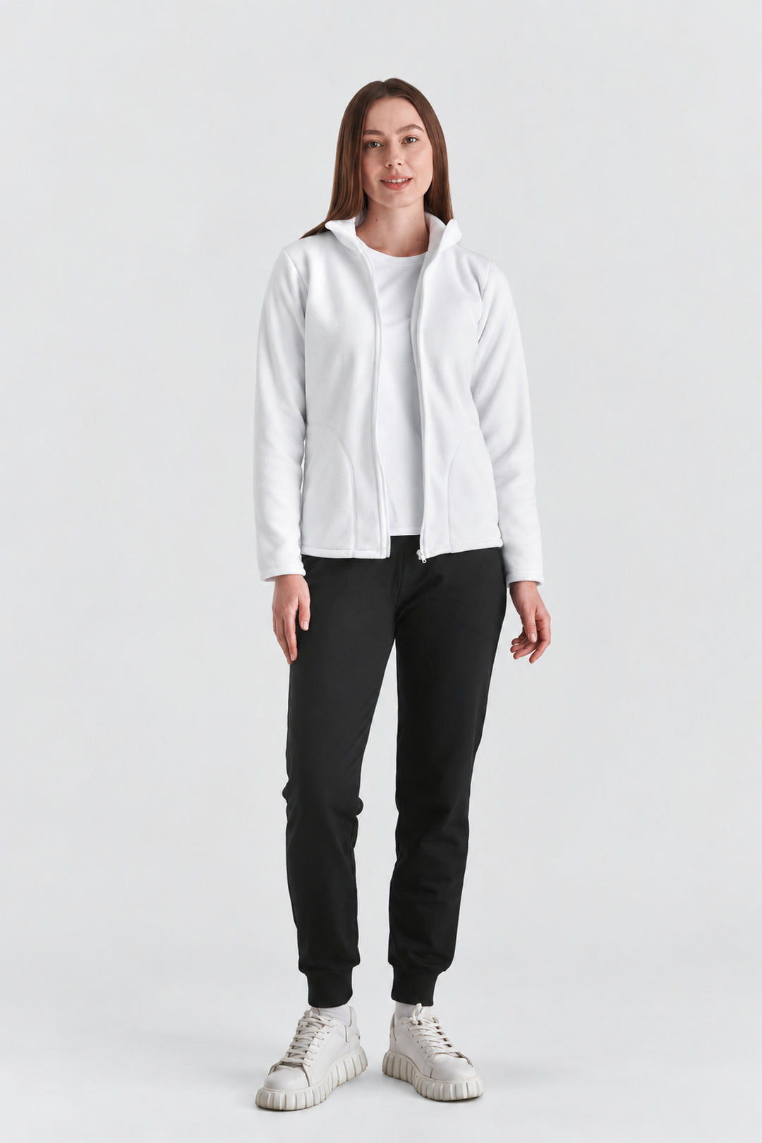 A woman modeling a white zip-up hoodie over a white top, paired with black jogger pants and white sneakers, against a plain light gray studio background.