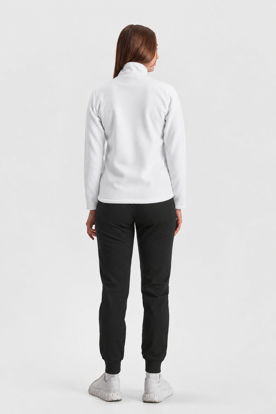 A woman viewed from the back wearing a white long-sleeve mock-neck top, black jogger pants, and white sneakers against a light gray studio background.
