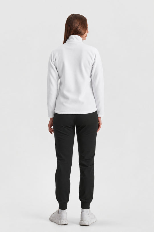 A woman viewed from the back wearing a white long-sleeve mock-neck top, black jogger pants, and white sneakers against a light gray studio background.
