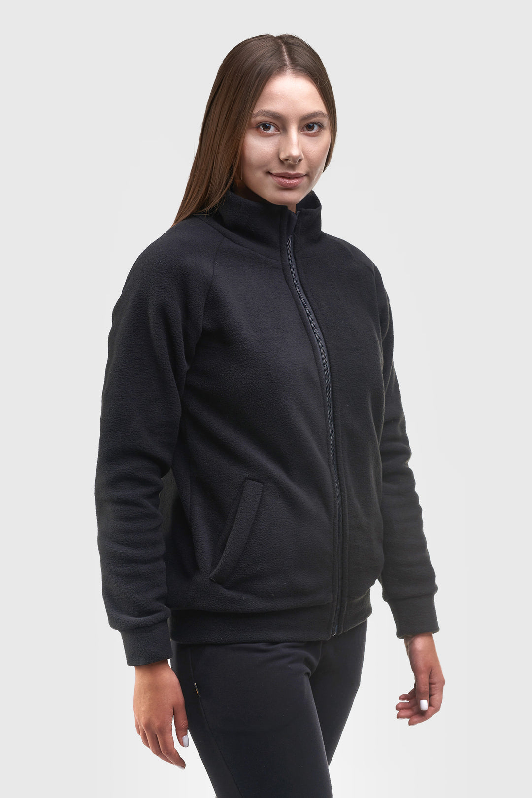 A woman modeling a black fleece zip-up jacket with a stand-up collar and side pockets, paired with black pants, against a light gray studio background.