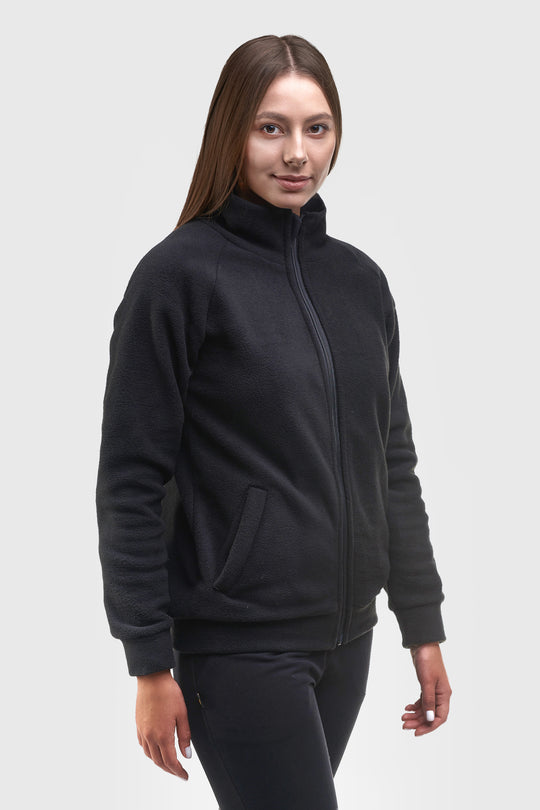 A woman modeling a black fleece zip-up jacket with a stand-up collar and side pockets, paired with black pants, against a light gray studio background.