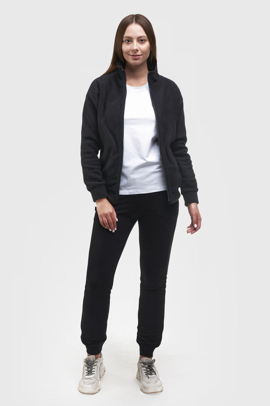 A woman modeling a black zip-up fleece jacket, white t-shirt, black jogger pants, and white sneakers against a light gray studio background.