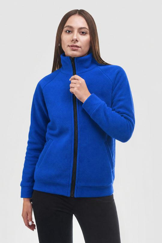 A woman wearing a bright royal blue fleece zip-up jacket with a black front zipper, paired with black pants, photographed against a light gray studio background.