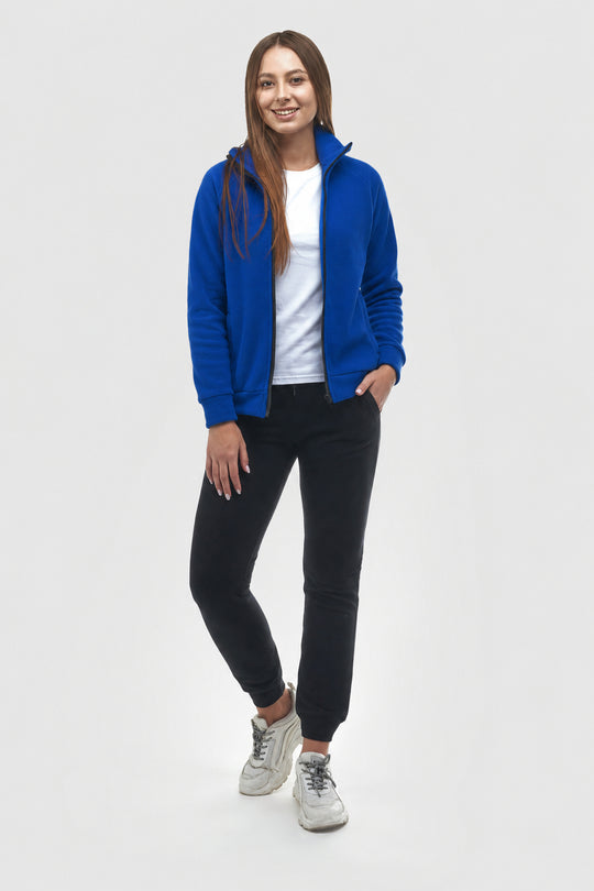 A woman modeling a bright royal blue zip-up fleece jacket over a white t-shirt, black jeans, and white sneakers, standing against a light gray studio background.