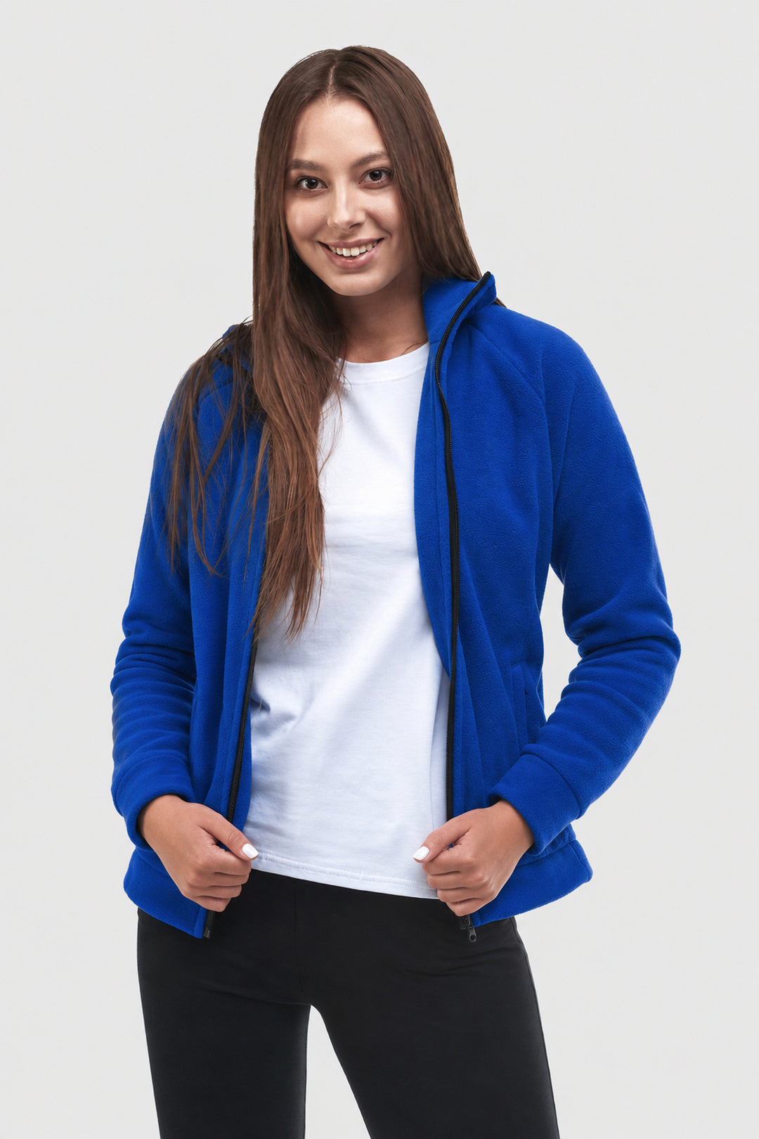 A woman modeling a bright royal blue zip-up hoodie with a black zipper, worn open over a white t-shirt and black pants, against a light gray studio background.