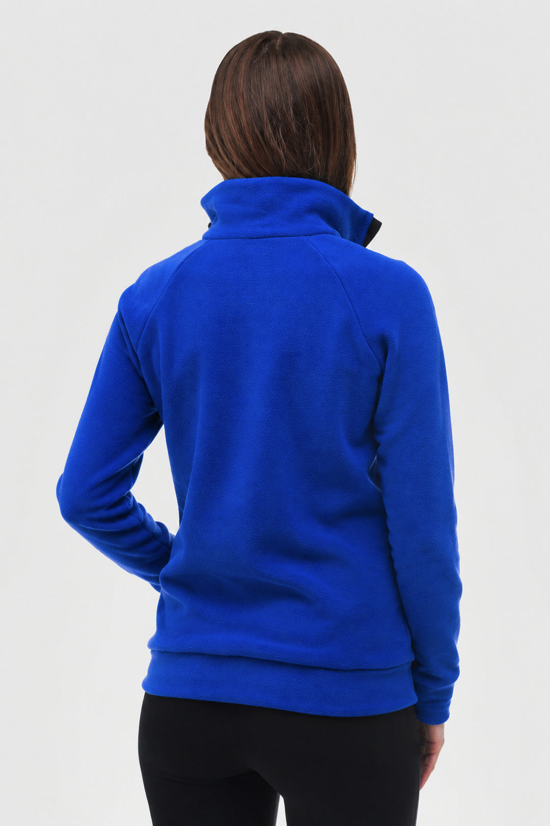 A woman viewed from the back wearing a bright royal blue fleece pullover with a high mock neck collar and black leggings, set against a plain light gray studio background.