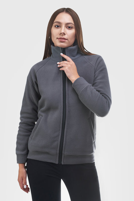 A woman wearing a dark gray zip-up fleece jacket with a high mock neck collar, paired with black pants, photographed against a solid light gray background.