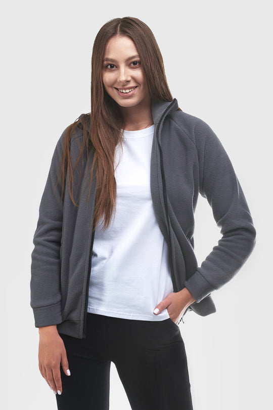 A woman modeling a dark gray zip-up hoodie over a white t-shirt and black pants, photographed against a solid light gray background.