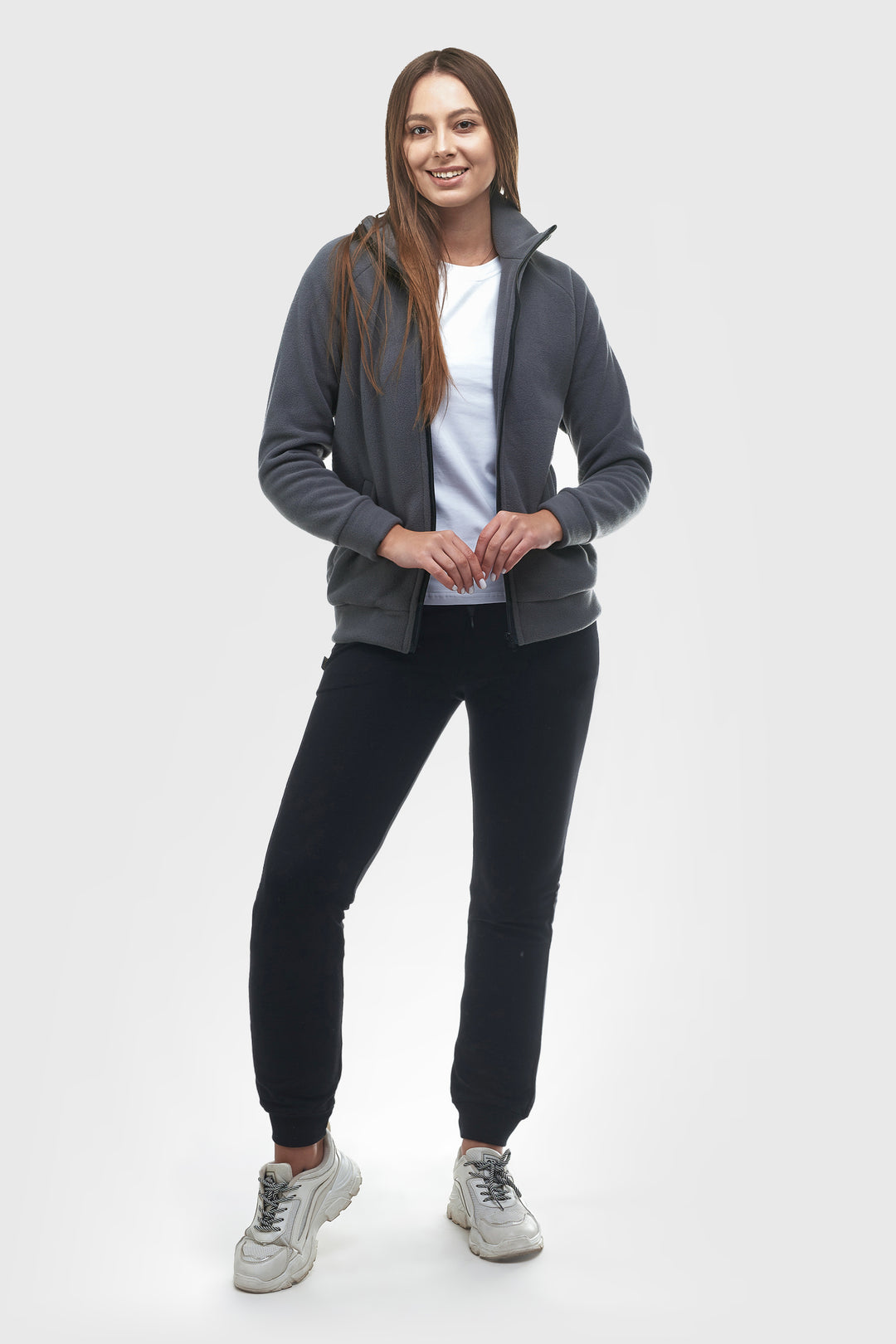 A woman modeling a dark gray zip-up hoodie over a white t-shirt, black joggers, and gray sneakers, standing against a light gray studio background.