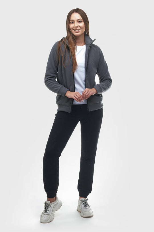A woman modeling a dark gray zip-up hoodie over a white t-shirt, black joggers, and gray sneakers, standing against a light gray studio background.
