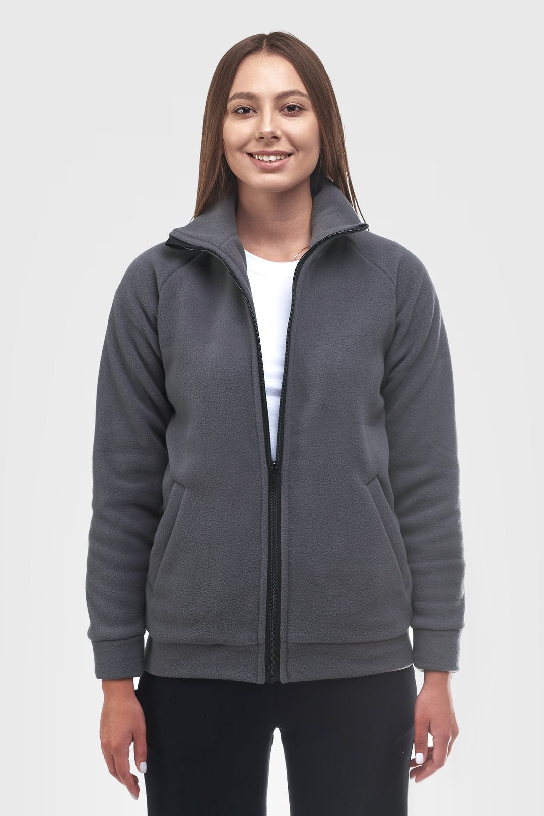 A woman modeling a dark gray zip-up fleece jacket with a stand-up collar, front pockets, and ribbed cuffs and hem, worn over a white t-shirt and black pants against a light gray studio background.