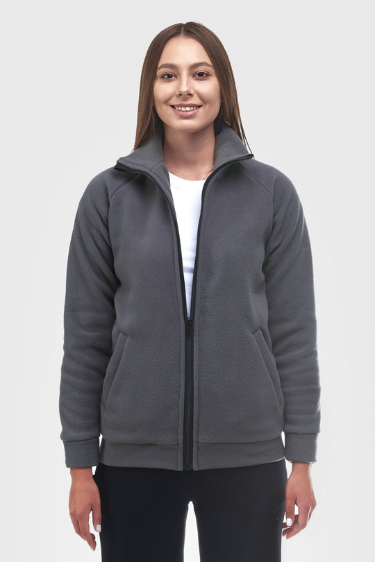 A woman modeling a dark gray zip-up fleece jacket with a stand-up collar, front pockets, and ribbed cuffs and hem, worn over a white t-shirt and black pants against a light gray studio background.