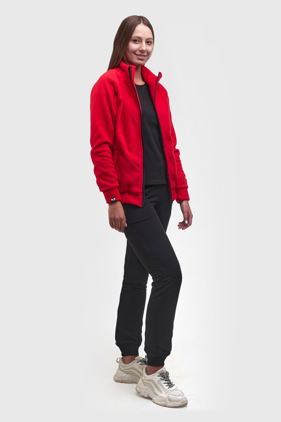 A woman modeling a bright red zip-up fleece jacket over a black top and black jogger pants, paired with chunky white sneakers, against a light gray studio background.