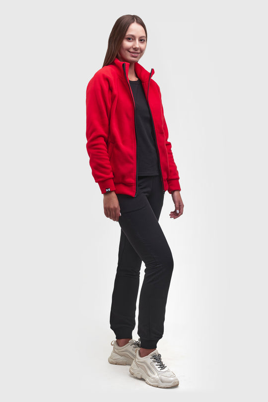 A woman modeling a bright red zip-up fleece jacket over a black top and black jogger pants, paired with chunky white sneakers, against a light gray studio background.