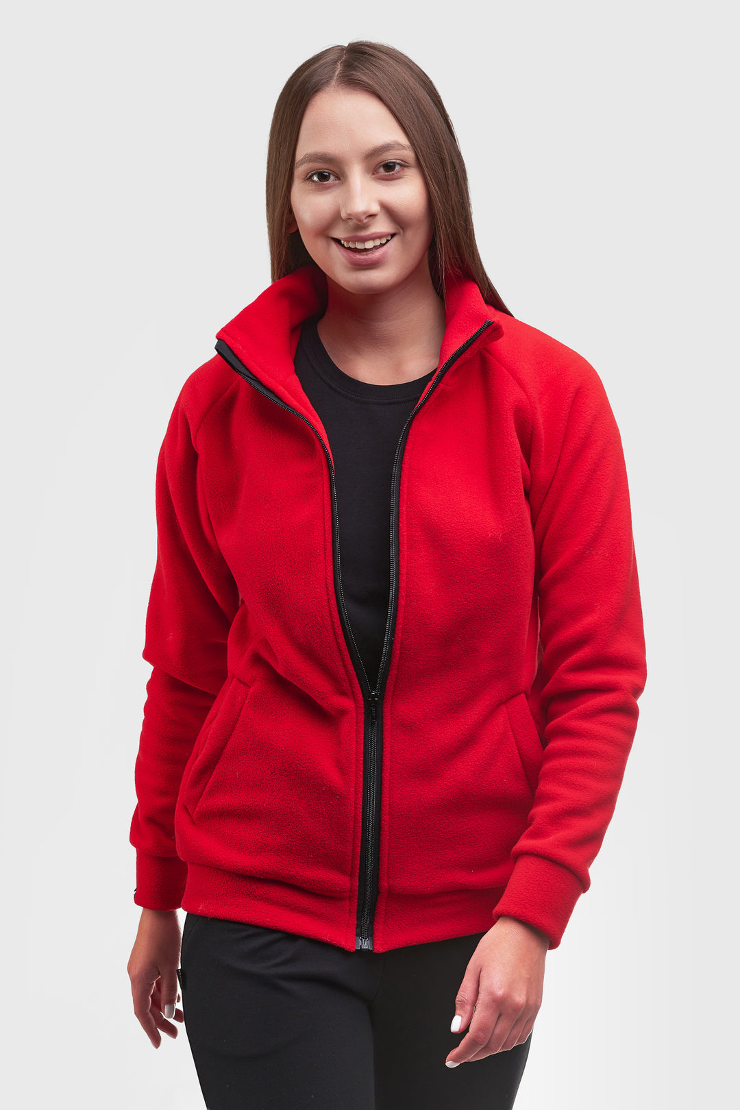 A woman wearing a bright red zip-up fleece jacket over a black top and black pants, standing against a light gray studio background.
