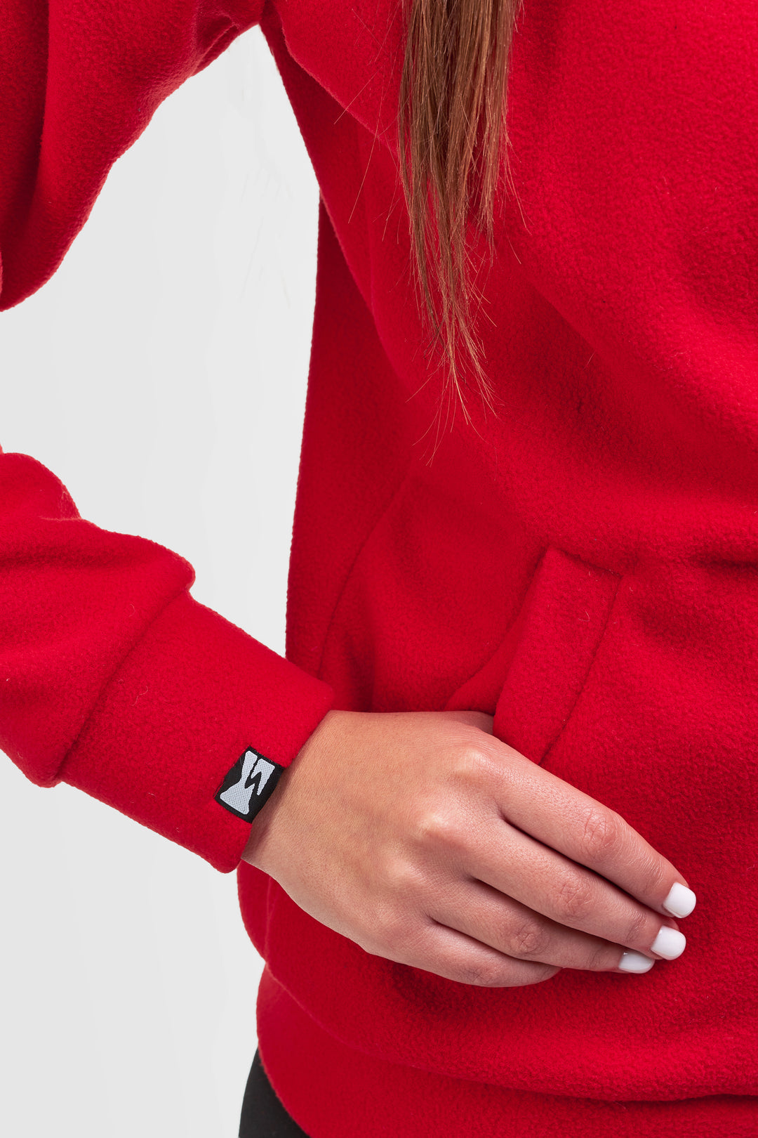 A close-up of a person wearing a bright red fleece hoodie, showing the sleeve cuff with a small black and white logo patch and the model's hand resting on the pocket.