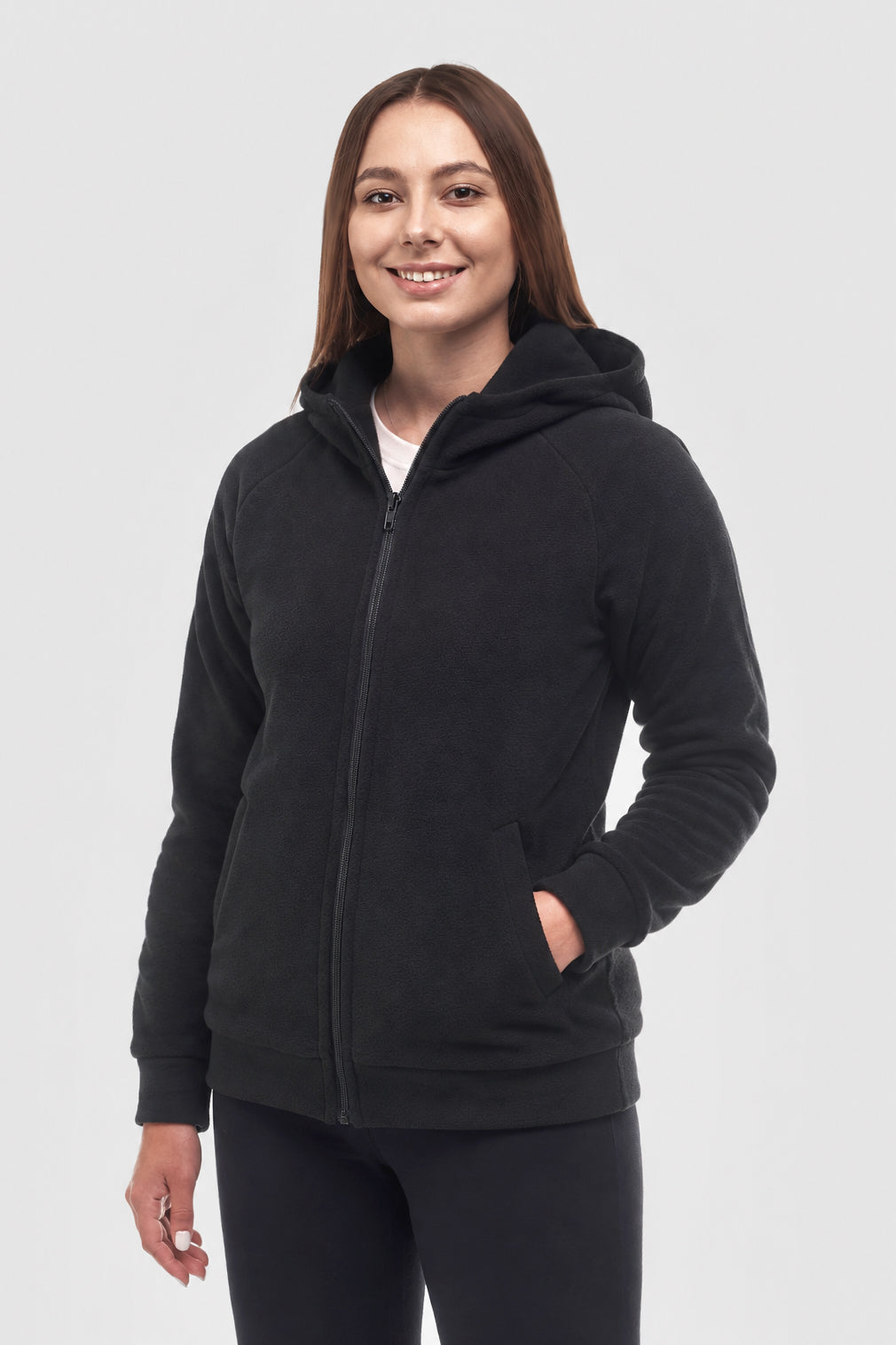 A woman wearing a black zip-up hoodie with a hood and front pockets, standing against a light gray studio background.