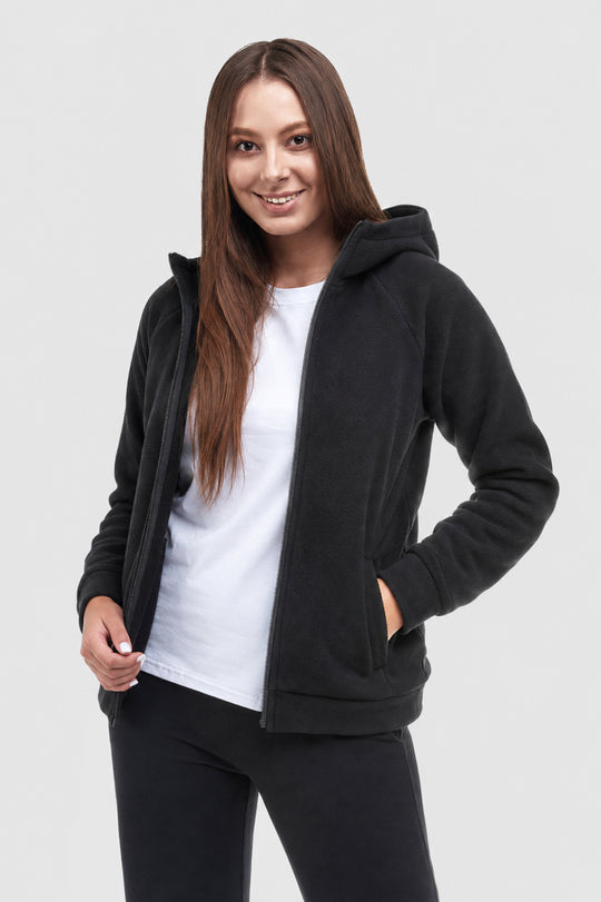 A woman modeling a black fleece zip-up hoodie over a white t-shirt and black pants, photographed against a solid light gray studio background.