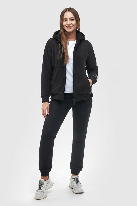 A woman modeling a black zip-up hoodie with a white t-shirt, black jogger pants, and white sneakers against a light gray studio background.