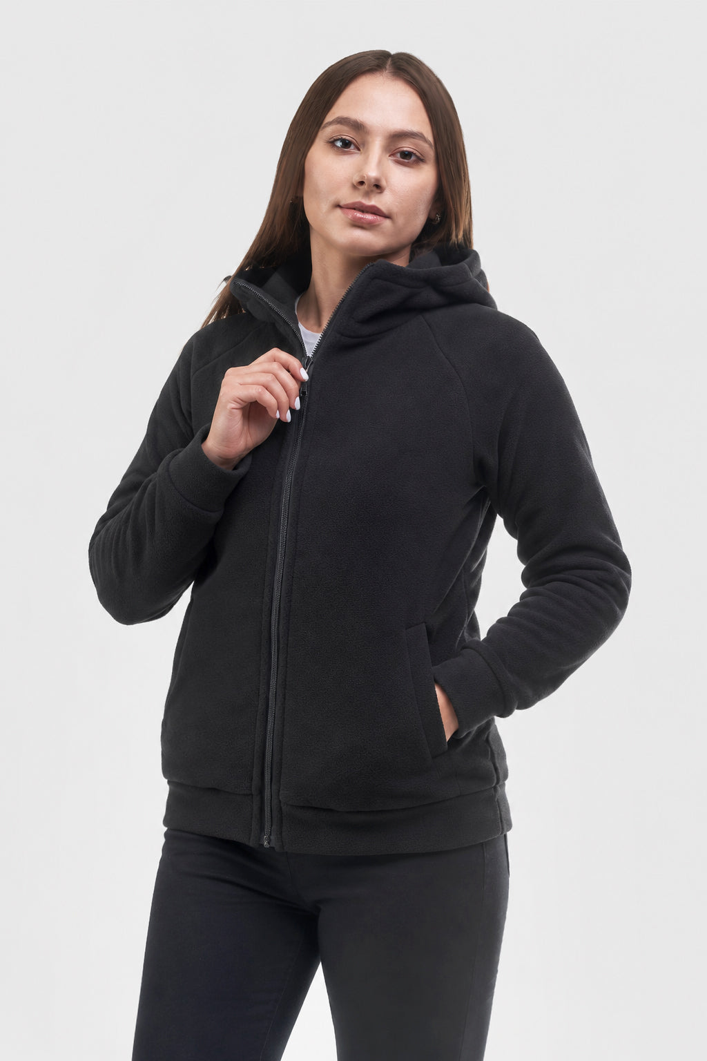A woman modeling a black zip-up hoodie with a hood and front pockets, paired with black leggings, against a light gray studio background.