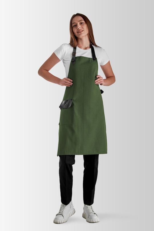 Woman wearing a green bib apron with black adjustable straps and flap pocket