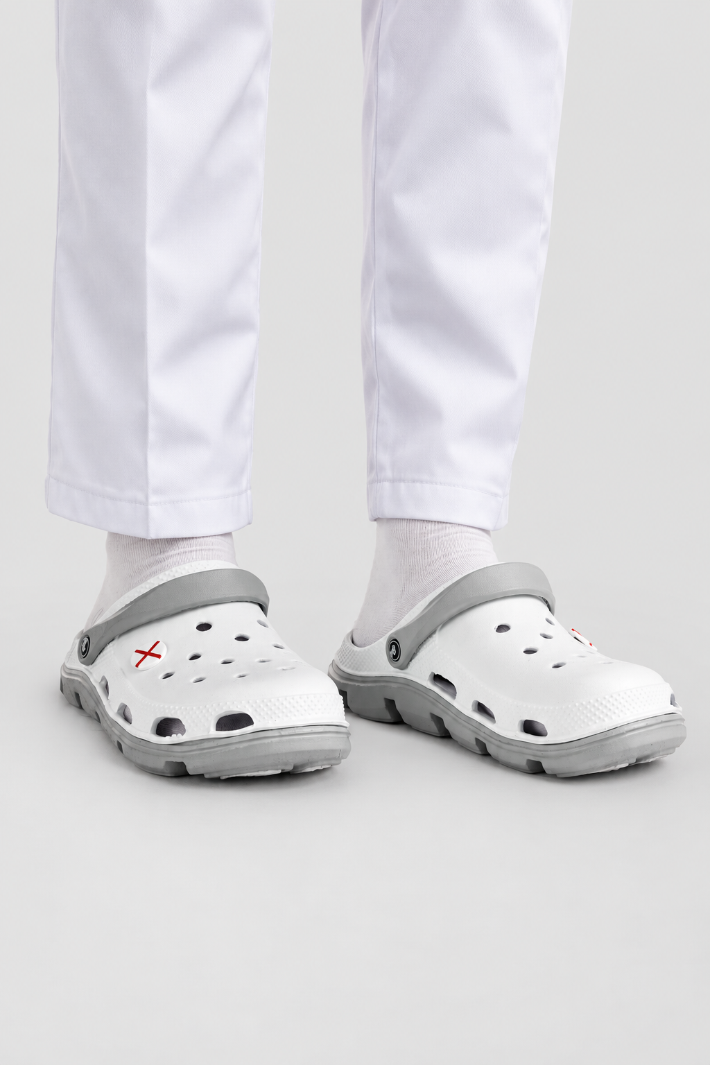 White and gray Croc-style clogs worn with white pants and socks, shown from the feet up against a light gray studio background.