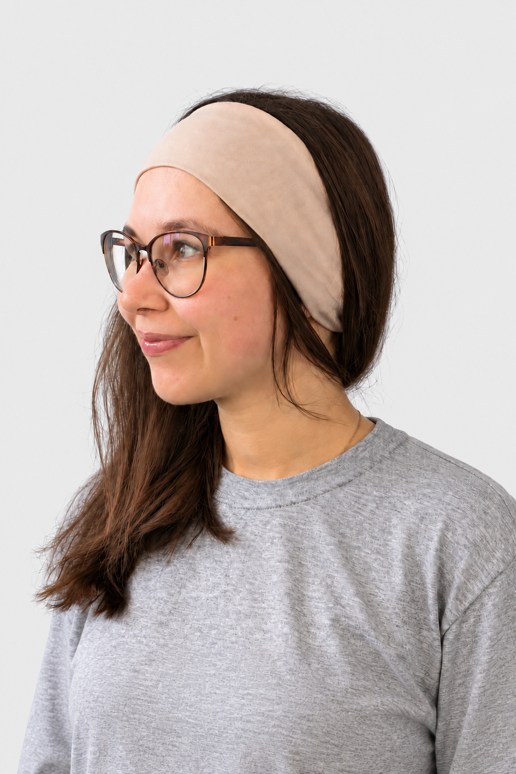 A woman with brown hair wearing a beige fabric headband and glasses, dressed in a gray t-shirt, shown in profile against a light gray background.